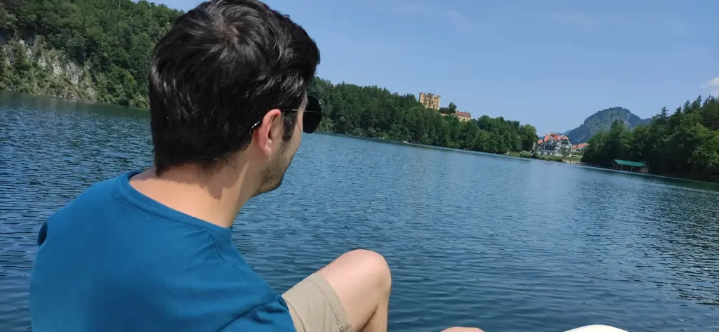 Ayal Othman sitting by a lake facing distant hills and a historic castle, symbolizing inspiration, travel, and creative growth.